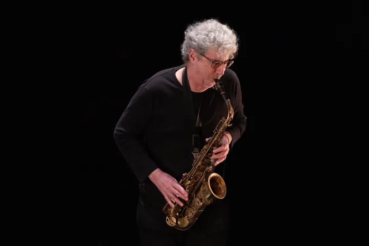 Person with glasses playing a saxophone on a dark stage.