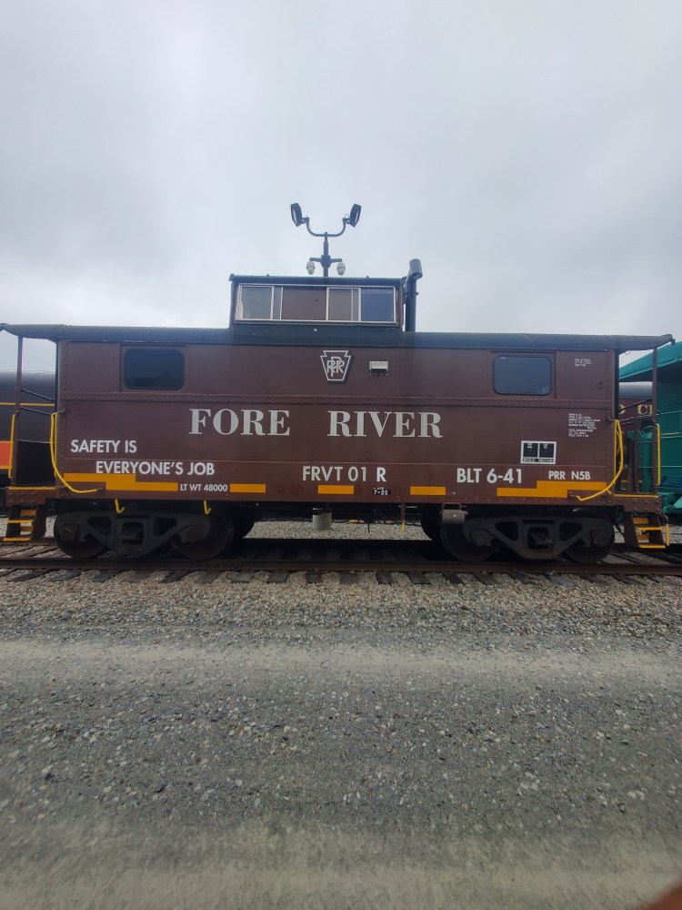Brown train caboose with 'Fore River' and safety sign on one side, parked on railway tracks.