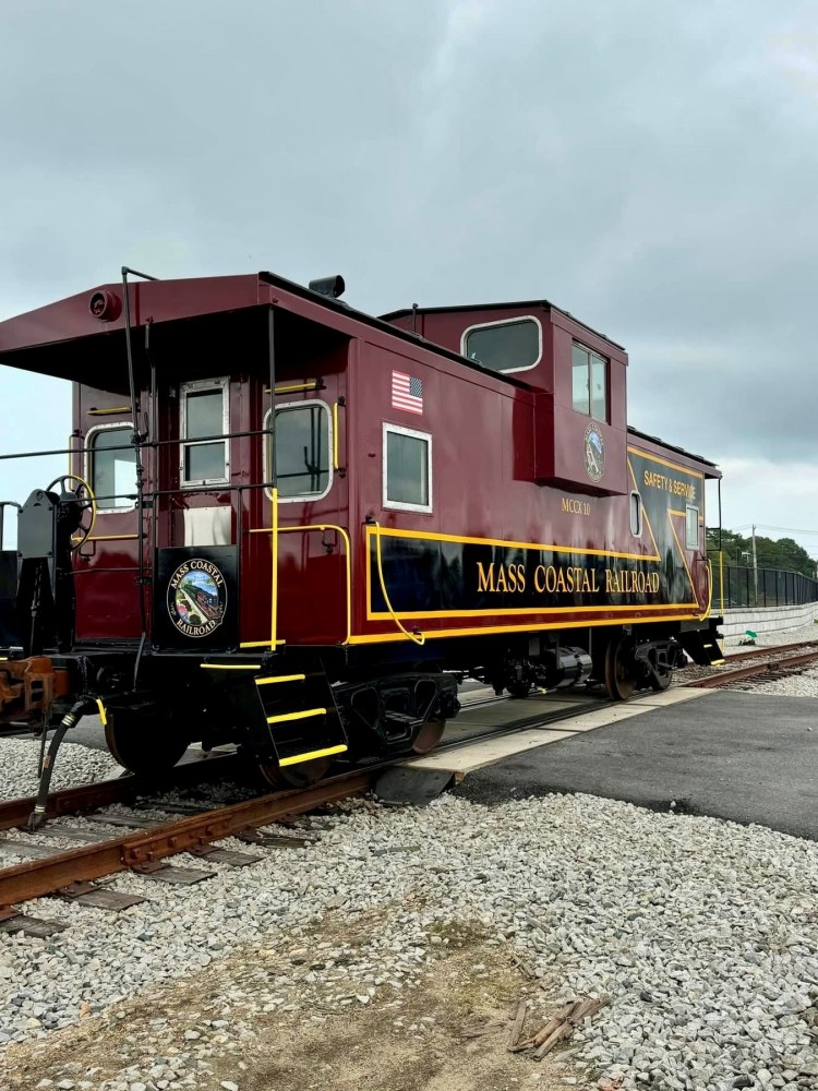 Red caboose labeled 'Mass Coastal Railroad' on train tracks under a cloudy sky.