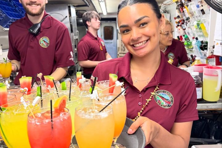 Smiling waitstaff holding a tray of colorful cocktails in a busy kitchen.