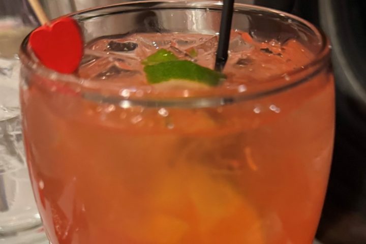 Red cocktail with ice, lime, and heart stirrer on a table.