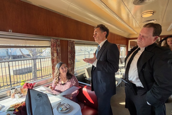 People dressed formally in a vintage train dining car, one seated at a table holding a menu.
