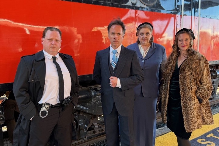 Four people in vintage clothing pose by a red and black train at a station.